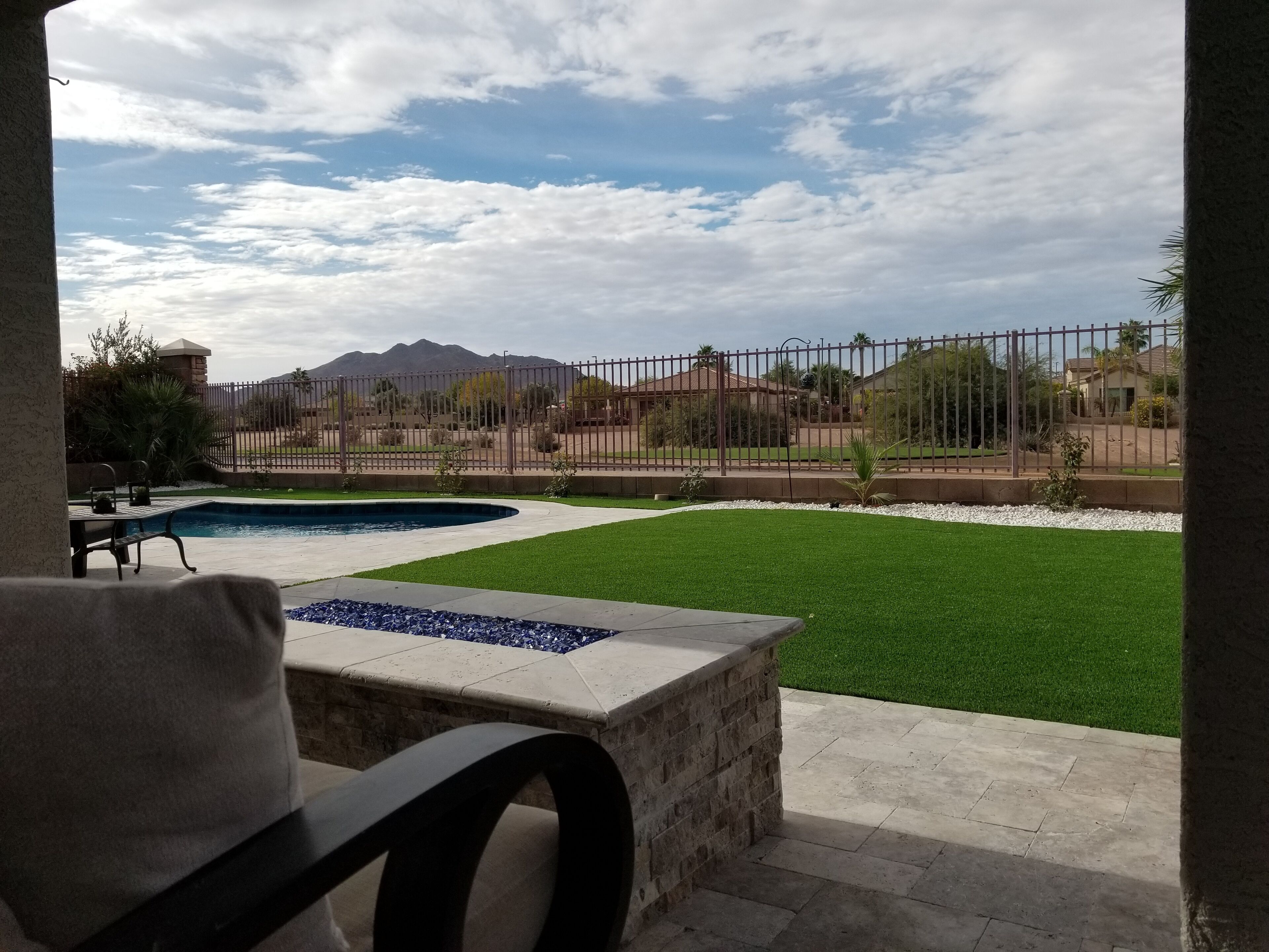 A pool area with a stone bench, a grassy lawn, and a view of a fenced property with mountains in the background.