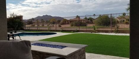 A pool area with a stone bench, a grassy lawn, and a view of a fenced property with mountains in the background.
