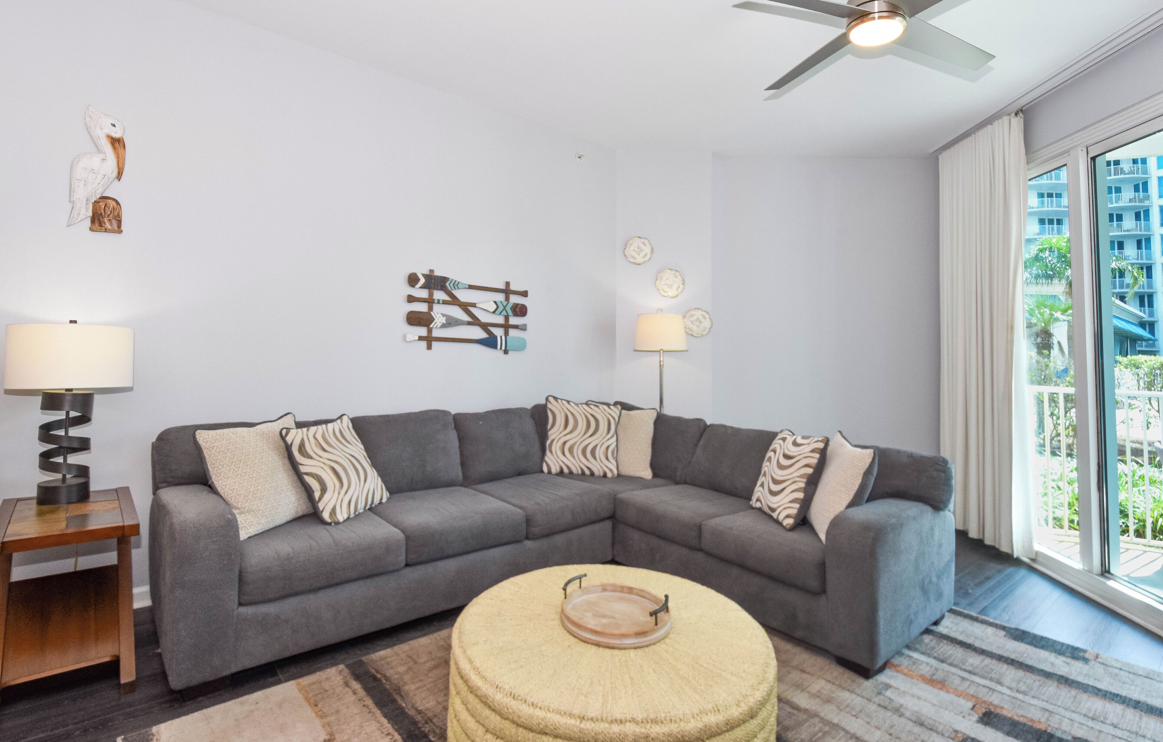 A modern living room with a grey sectional sofa, a small round ottoman, a floor lamp, and a ceiling fan.