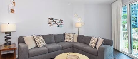 A modern living room with a grey sectional sofa, a small round ottoman, a floor lamp, and a ceiling fan.