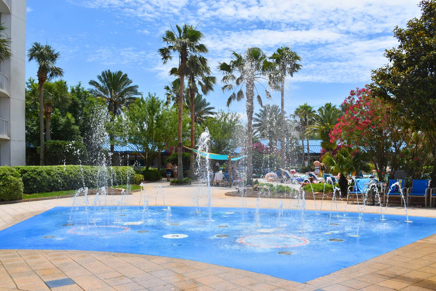 Une fontaine avec plusieurs jets d’eau, entourée de palmiers et bordée d’une piscine.