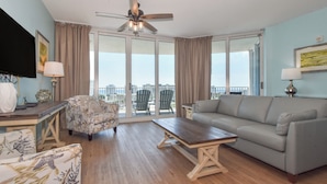 Een ruime woonkamer met een grijze bank, een houten salontafel, een fauteuil met een patroon en een plafondventilator.