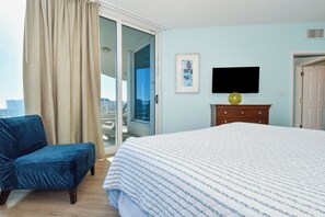 Un dormitorio con cama, una silla azul, cómoda, televisor y puerta que da a un balcón.