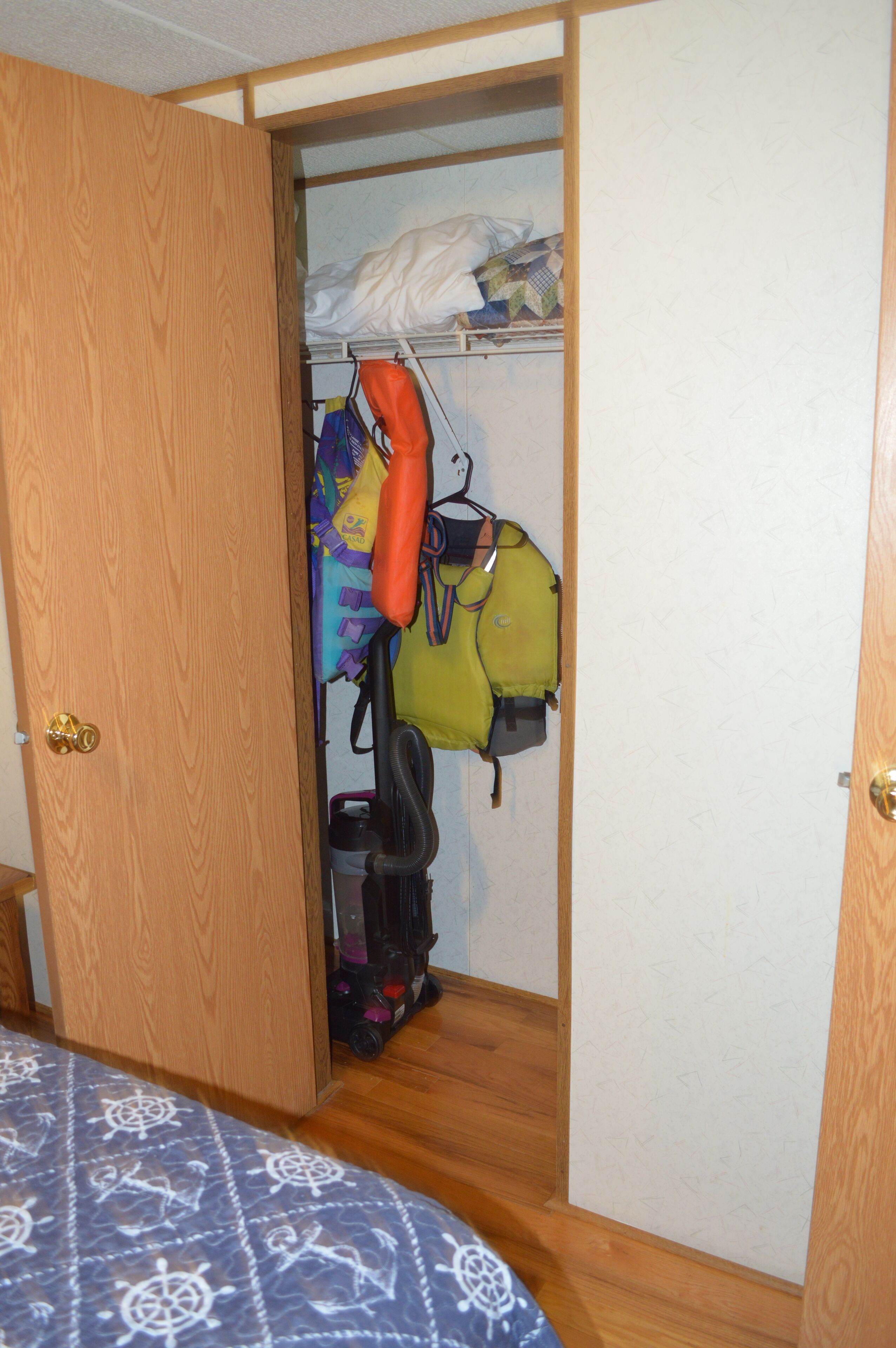 A bedroom with a bed, a wooden door, and a closet containing a hanging rack with bags and a vacuum cleaner.