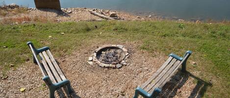 A lakeside setting with two wooden benches facing a fire pit.
