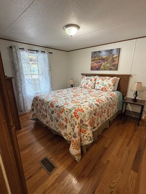 Un dormitorio con colcha estampada, cabecera de madera, mesita de noche con lámpara y ventana con cortinas traslúcidas.