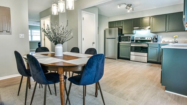 A modern kitchen with stainless steel appliances, a dining area with a round table and blue chairs, and a living area with a large window. - Roomy 3 BR Condo Walkable to IU (Bloomington)