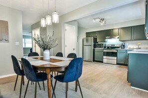 Una cocina moderna con electrodomésticos de acero inoxidable, un comedor con una mesa redonda y sillas azules, y una sala de estar con un ventanal grande.