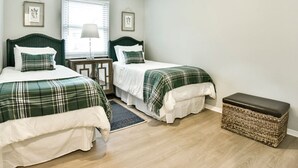 A bedroom with two beds, a nightstand, a lamp, a window with blinds, and a woven bench. - Roomy 3 BR Condo Walkable to IU (Bloomington)