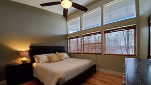 A bedroom with a bed, a nightstand, a lamp, a ceiling fan, and large windows with blinds. - Serene Lake Home (Fort Scott)