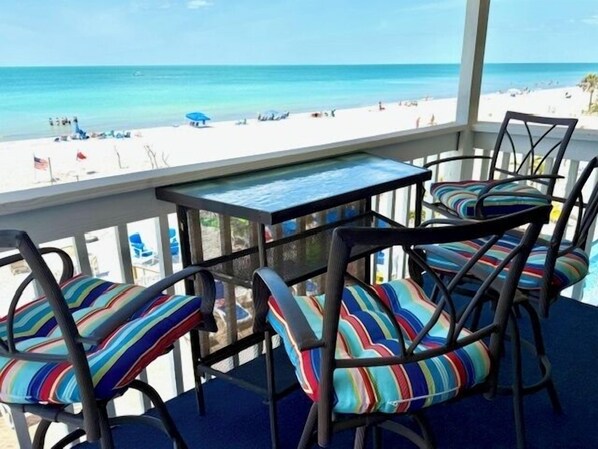 A covered outdoor seating area with a table and chairs, overlooking a beach with people and umbrellas.