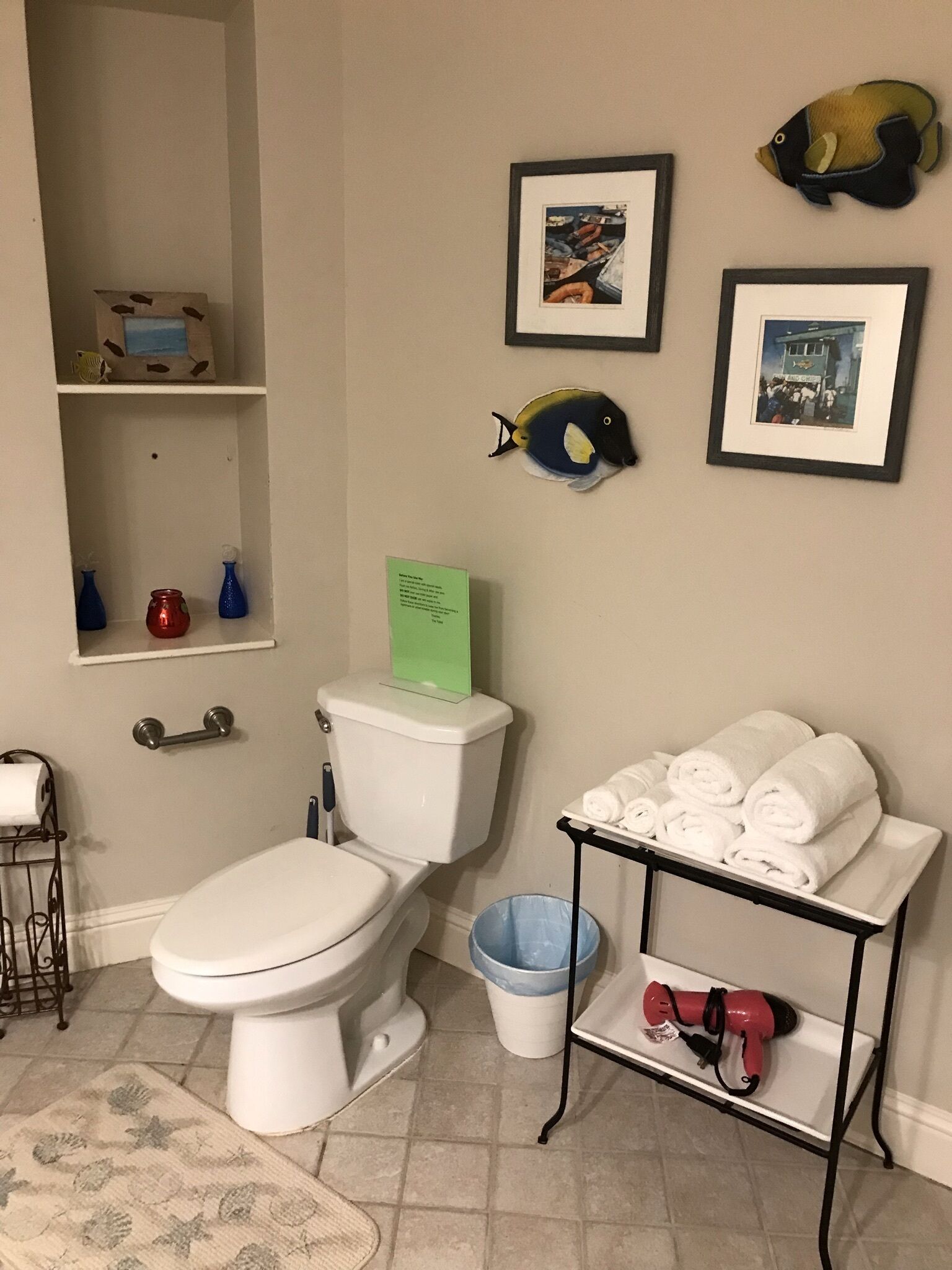 Une salle de bain comprenant des toilettes, une étagère avec des objets décoratifs, un tableau encadré et un porte-serviettes.