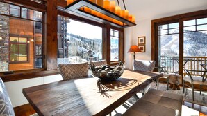 A dining area with a wooden table, a bowl of fruits, and a view of a snowy mountain landscape through large windows.