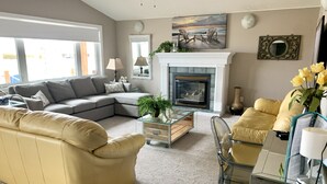 A living room with a sectional sofa, a glass coffee table, a fireplace, and a painting of chairs on a beach.