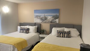 Zwei Einzelbetten mit weißen Laken und gelben Tagesdecken, ein Nachttisch mit Lampe sowie ein Bild einer Strandansicht an der Wand.