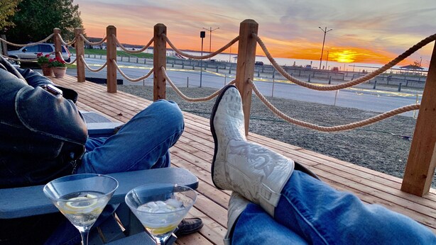 A wooden deck with a view of the sunset over the water, a rope barrier, and a table with a drink. - 201 - Executive Rm Lake View- Balcony 2 DBL (Port Elgin)