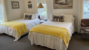 A hotel room with two beds, each with a yellow throw blanket, white bedding, and decorative pillows. There is a painting of a boat on the wall, a small table with a lamp, and a chair. - 202 - Executive Rm Lake View- Balcony 2 DBL (Port Elgin)