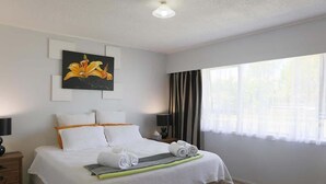 A neatly made bed with white linens and a green accent, a painting of a flower on the wall, a nightstand with a lamp, and a large window with sheer curtains.