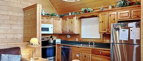 A cozy kitchen with wooden cabinets, a stainless steel refrigerator, a microwave, and a sink. There is a dining table with chairs and a couch with a patterned cushion.