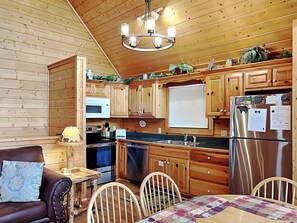 A cozy kitchen with wooden cabinets, a stainless steel refrigerator, a microwave, and a sink. There is a dining table with chairs and a couch with a patterned cushion. - Trail's End by Jackson Mountain Rentals (Gatlinburg)
