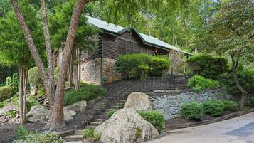 A log cabin with a stone foundation, surrounded by lush greenery and a stone staircase leading to the entrance.