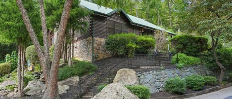 A log cabin with a stone foundation, surrounded by lush greenery and a stone staircase leading to the entrance.