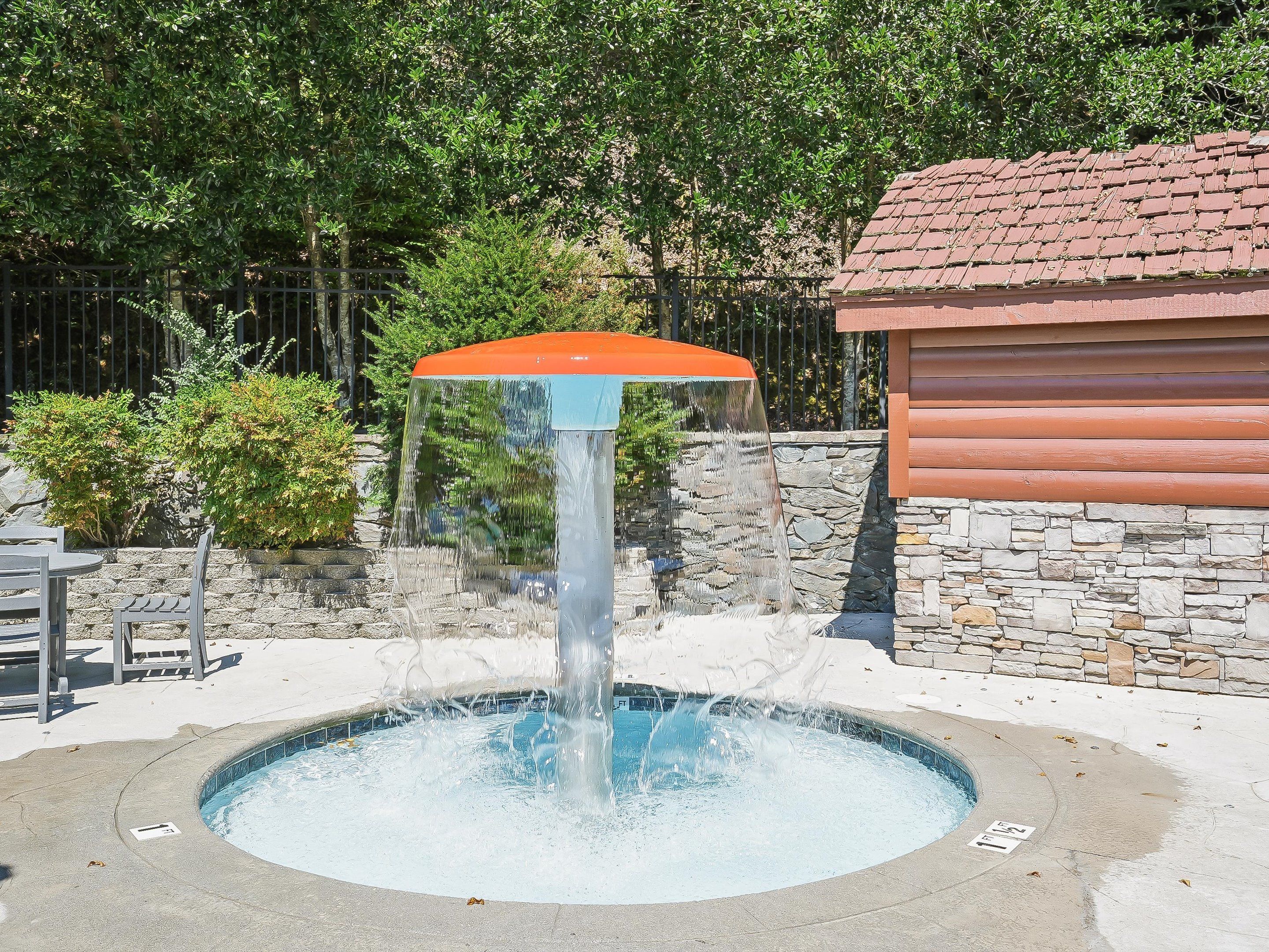 A circular pool with a central fountain, surrounded by a stone wall and a wooden building.