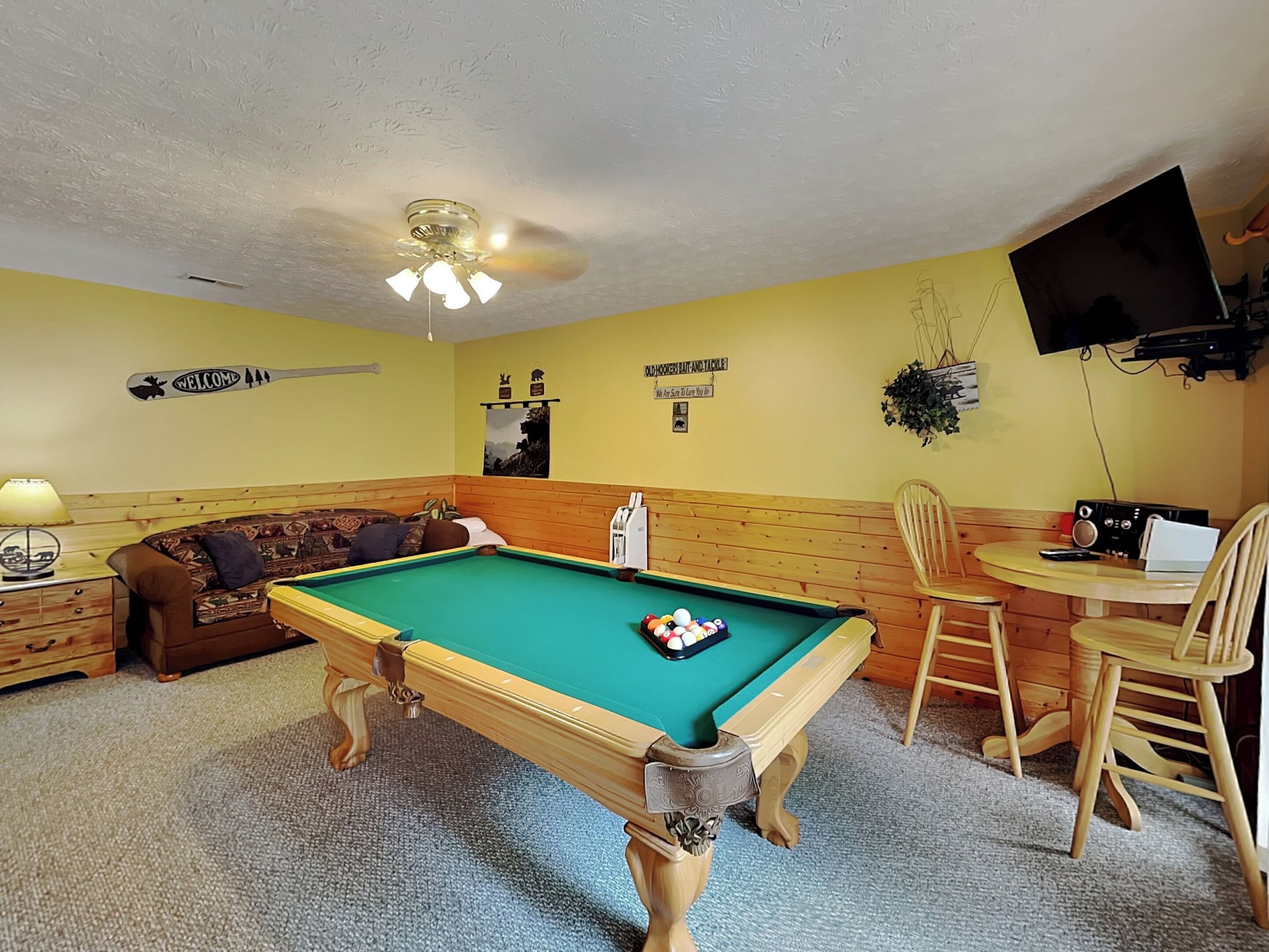 Une table de billard avec des billes, un canapé, un téléviseur et une chaise dans une pièce aux murs en bois et dotée d’un ventilateur de plafond.