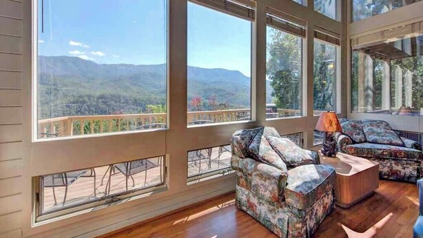 Une pièce spacieuse avec de grandes fenêtres donnant sur les montagnes, une terrasse en bois à l’extérieur et un coin salon confortable avec des canapés à motifs.