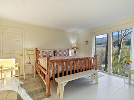 A bedroom with a wooden bed, a bench, a nightstand, a lamp, and a view of the outdoors.