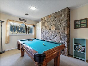 A pool table in a room with a stone wall, a window with a view of trees, and a billiards sign. - Shooting Star by Jackson Mountain Rentals (Gatlinburg)