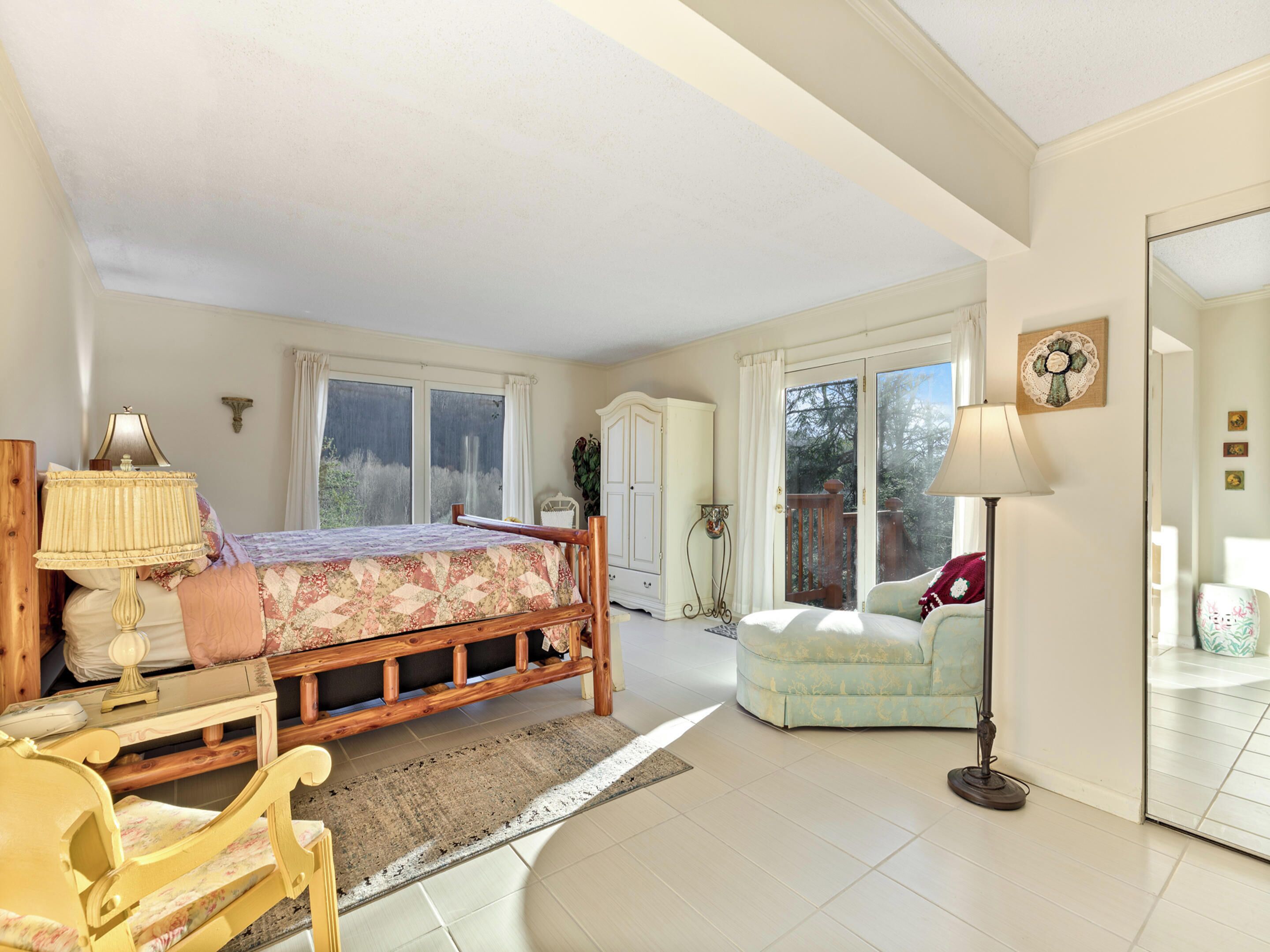 A spacious bedroom with a large bed, a wooden nightstand, a desk, a chair, and a floor lamp. There is a wardrobe, a window with curtains, and a door leading to a balcony.