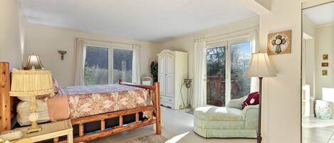 A spacious bedroom with a large bed, a wooden nightstand, a desk, a chair, and a floor lamp. There is a wardrobe, a window with curtains, and a door leading to a balcony.