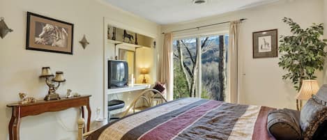 Ein Schlafzimmer mit einem großen Bett, einem Schreibtisch mit Fernseher, einem Spiegel und Blick auf Bäume durch ein Fenster.