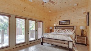 Une chambre à coucher située dans une cabane en bois, comprenant un lit, deux tables de chevet, un ventilateur de plafond et de grandes fenêtres équipées de stores.