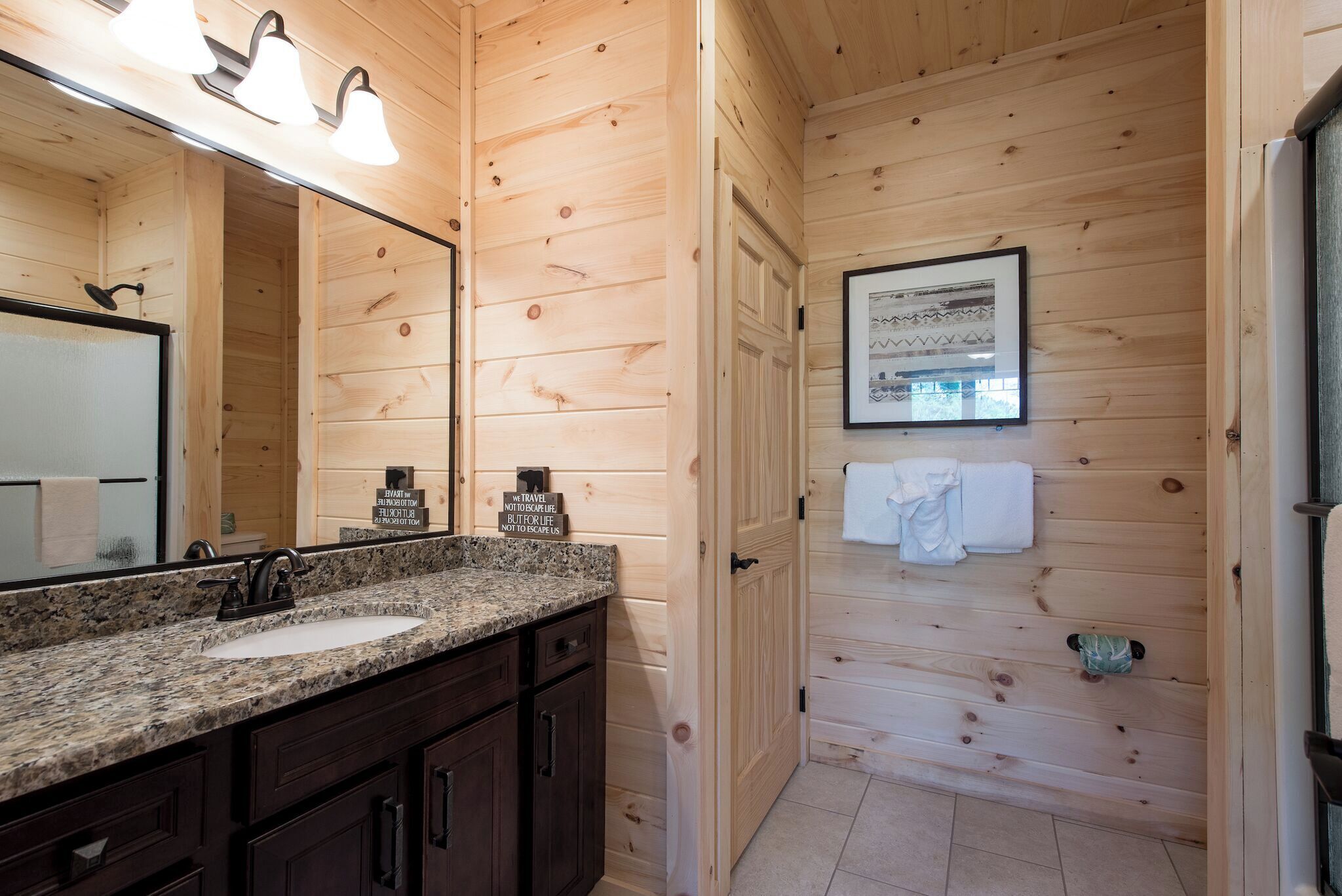 A wooden cabin bathroom with a granite countertop sink, dark wooden cabinets, a large mirror, and a framed picture on the wall.
