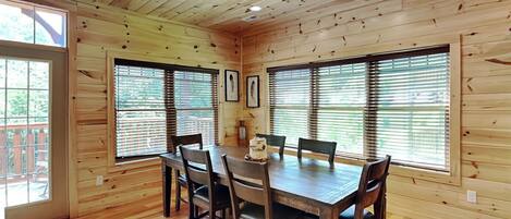 A wooden cabin with a dining area featuring a large table and chairs, large windows with blinds, and a wooden floor.