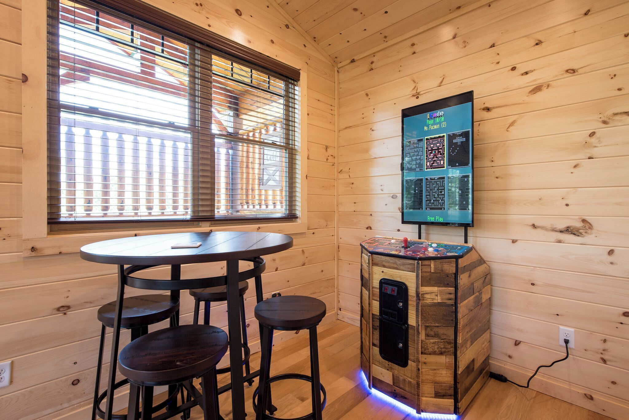 A wooden interior with a round table, stools, a jukebox, and a wall-mounted screen displaying a QR code and text.