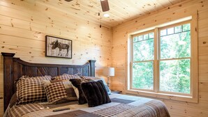 A wooden cabin bedroom with a large bed, a mounted picture of a moose, and a window with a forest view. - Smoky Haven by Jackson Mountain Rentals (Gatlinburg)