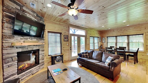 A cozy cabin interior with a stone fireplace, a leather sofa, a dining table, and a ceiling fan. - Smoky Haven by Jackson Mountain Rentals (Gatlinburg)