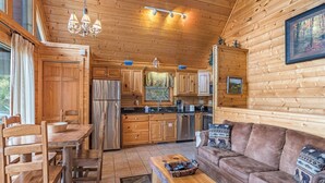 A cozy cabin interior with wooden walls, a dining area with a wooden table and chairs, a sofa, and a kitchen with wooden cabinets and appliances. - Saddle Ridge by Jackson Mountain Rentals (Gatlinburg)