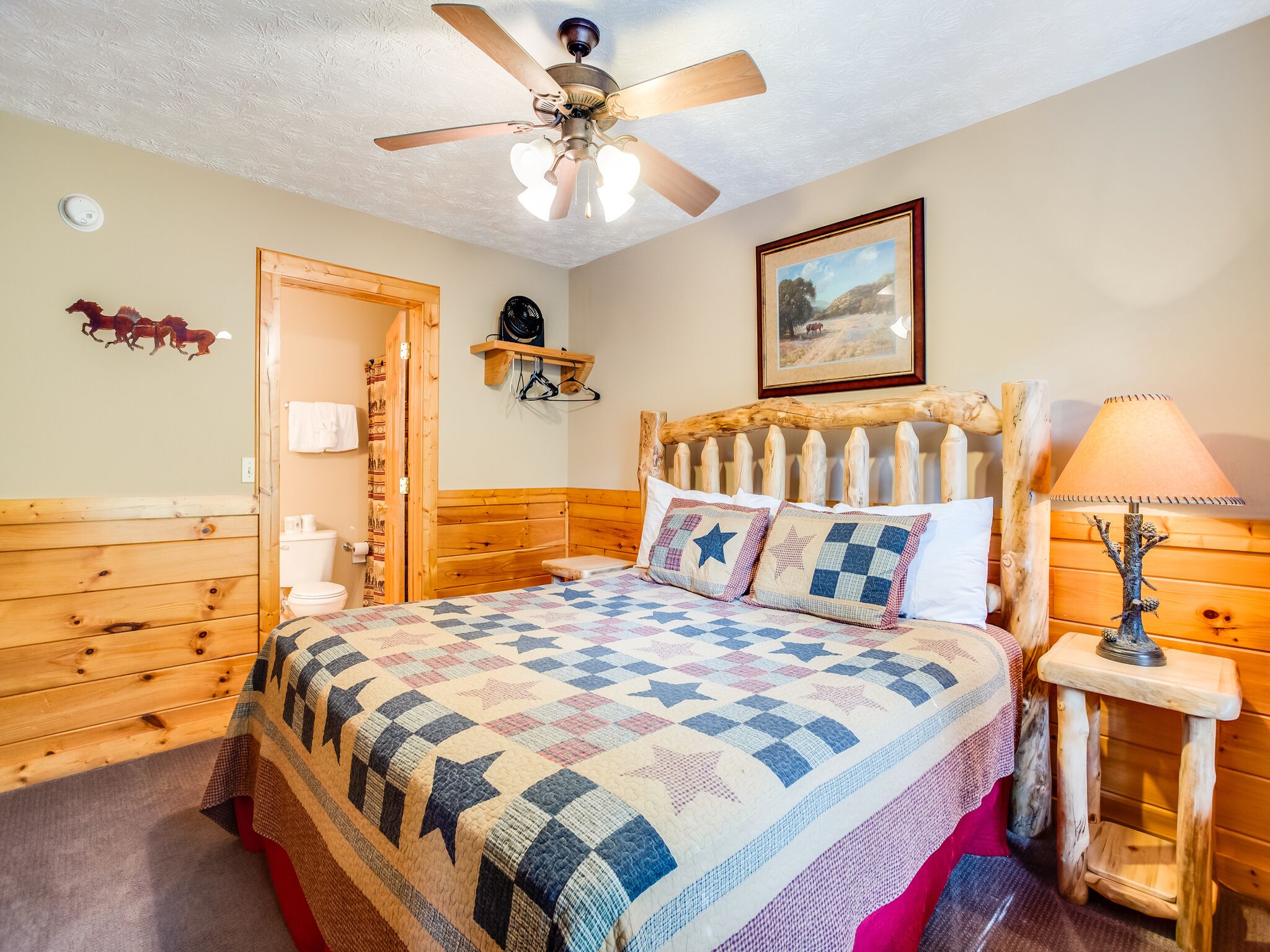 A cozy room with a wooden bed, a ceiling fan, a framed picture on the wall, and a small table with a lamp.
