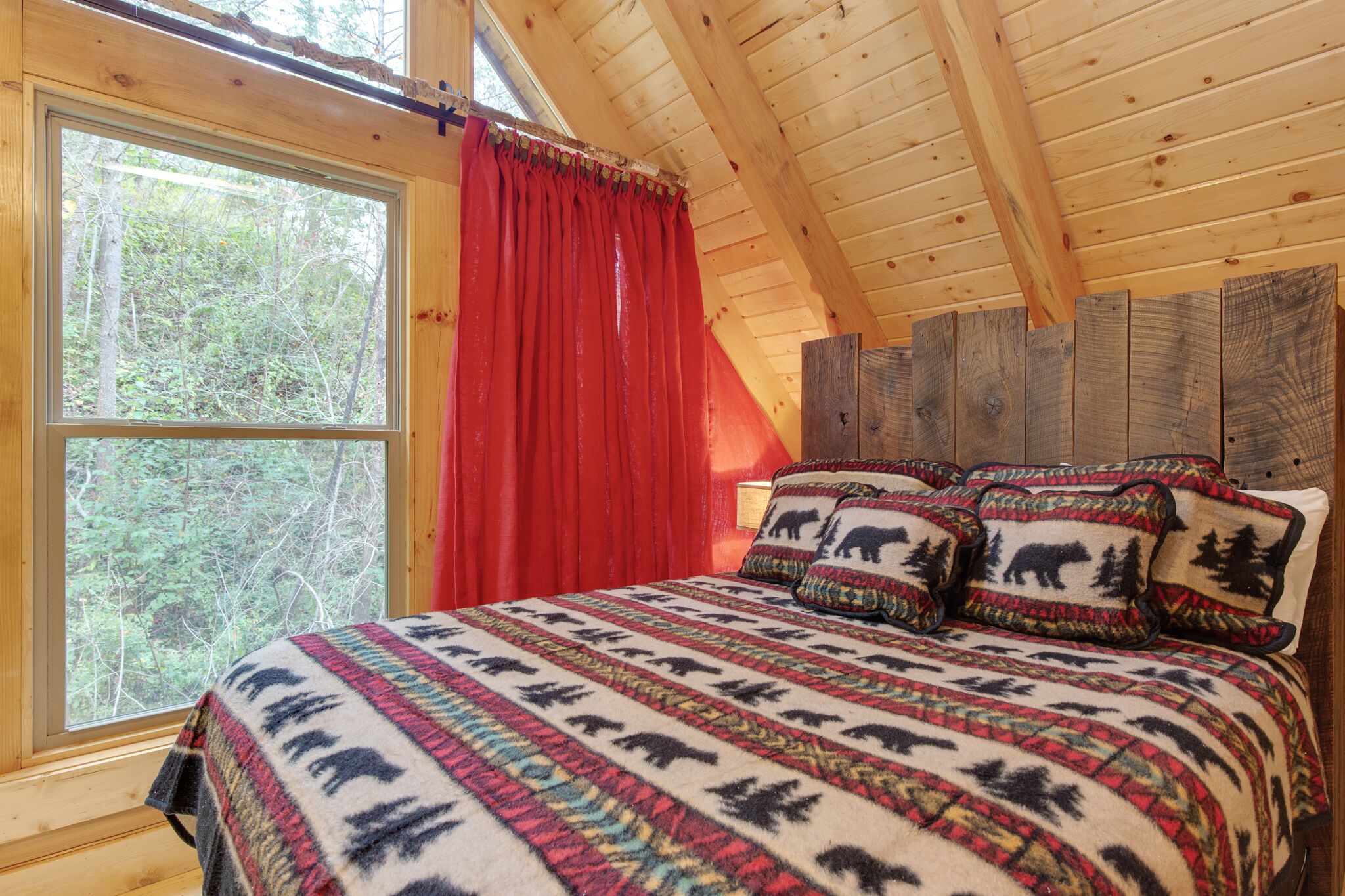 Un dormitorio de cabaña de madera con un ventanal grande, cortinas rojas, una cama con ropa de cama estampada y almohadones con motivos de animales.