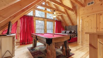 Une pièce mansardée chaleureuse, avec une table de billard, des rideaux rouges et un plafond en pente orné de poutres en bois.