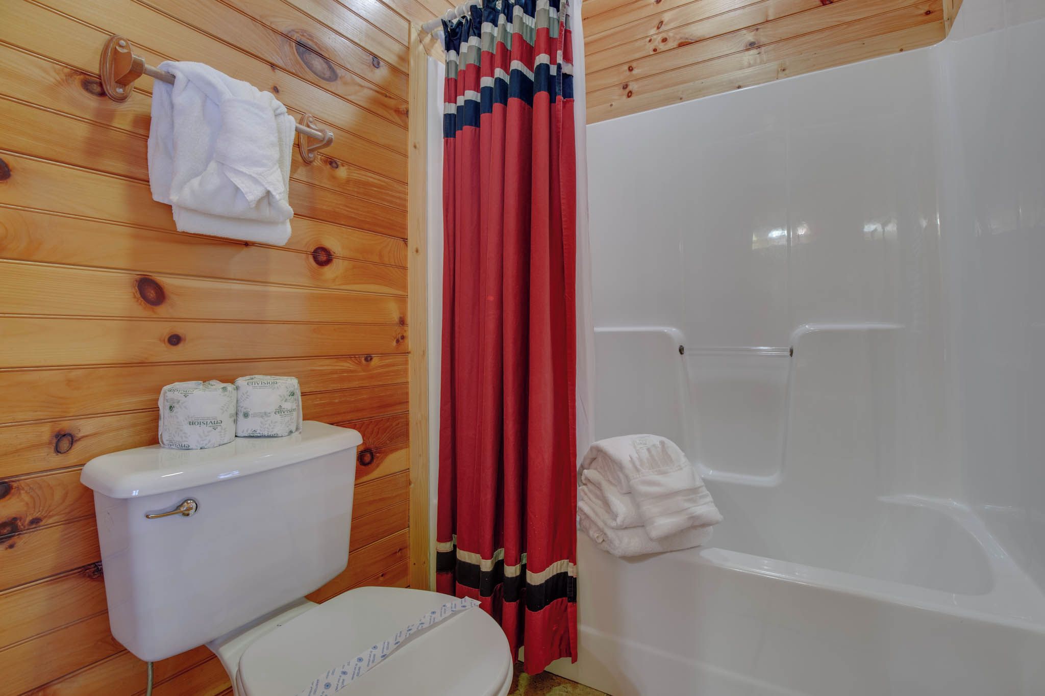 A wooden-paneled bathroom with a toilet, two rolls of toilet paper, a white bathtub, and a red and black striped shower curtain.