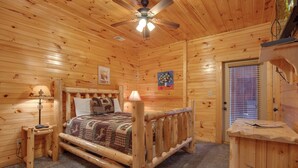 Un dormitorio de cabaña de madera con cama, mesita de noche, lámpara, ventilador de techo y ventana con persianas.