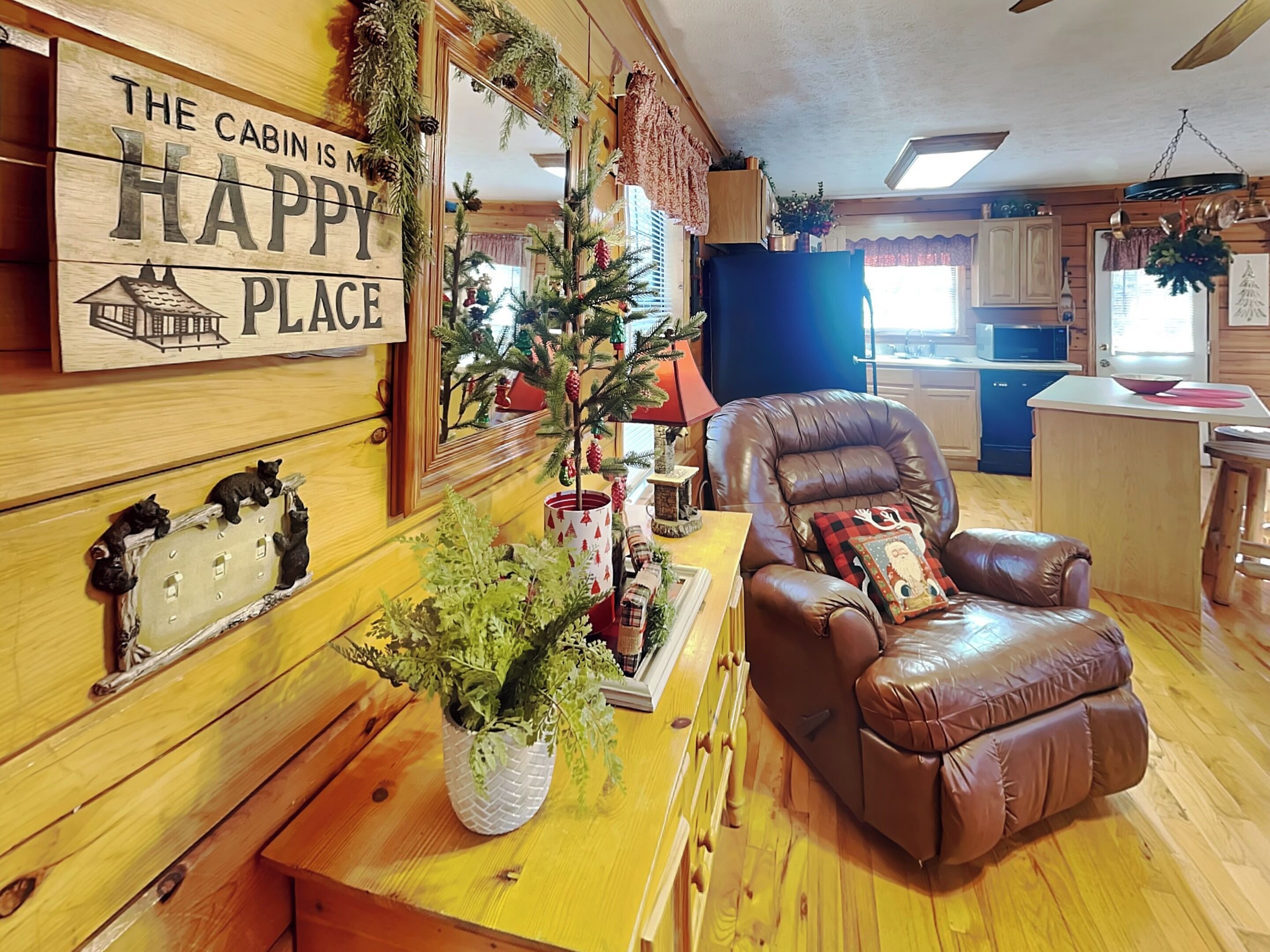 Een gezellig interieur van een chalet met een houten eettafel, een leren bank, een kerstboom en een bordje met de tekst 'THE CABIN IS A HAPPY PLACE'.