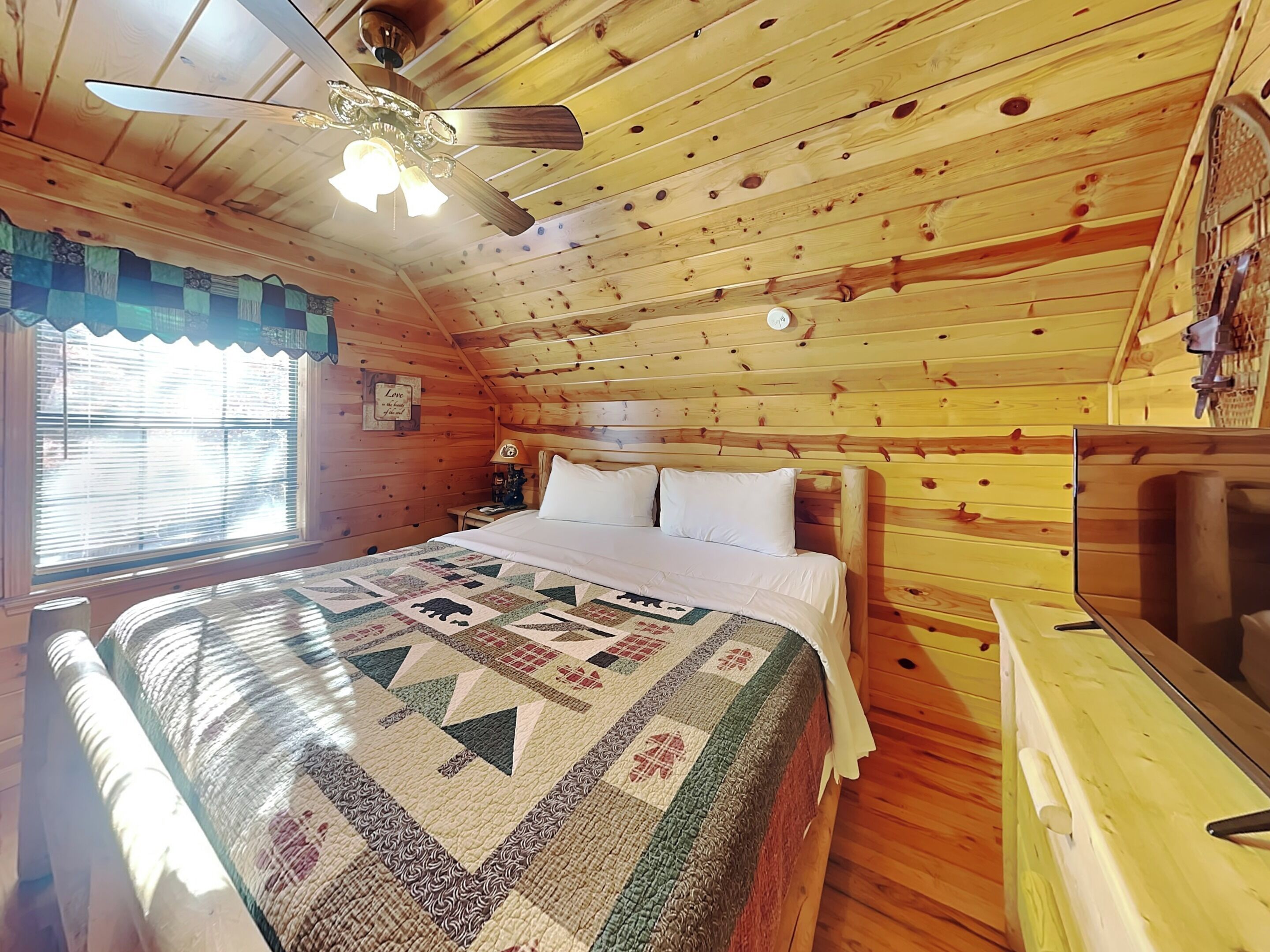 A cozy wooden cabin bedroom with a bed, a nightstand, a lamp, and a ceiling fan.