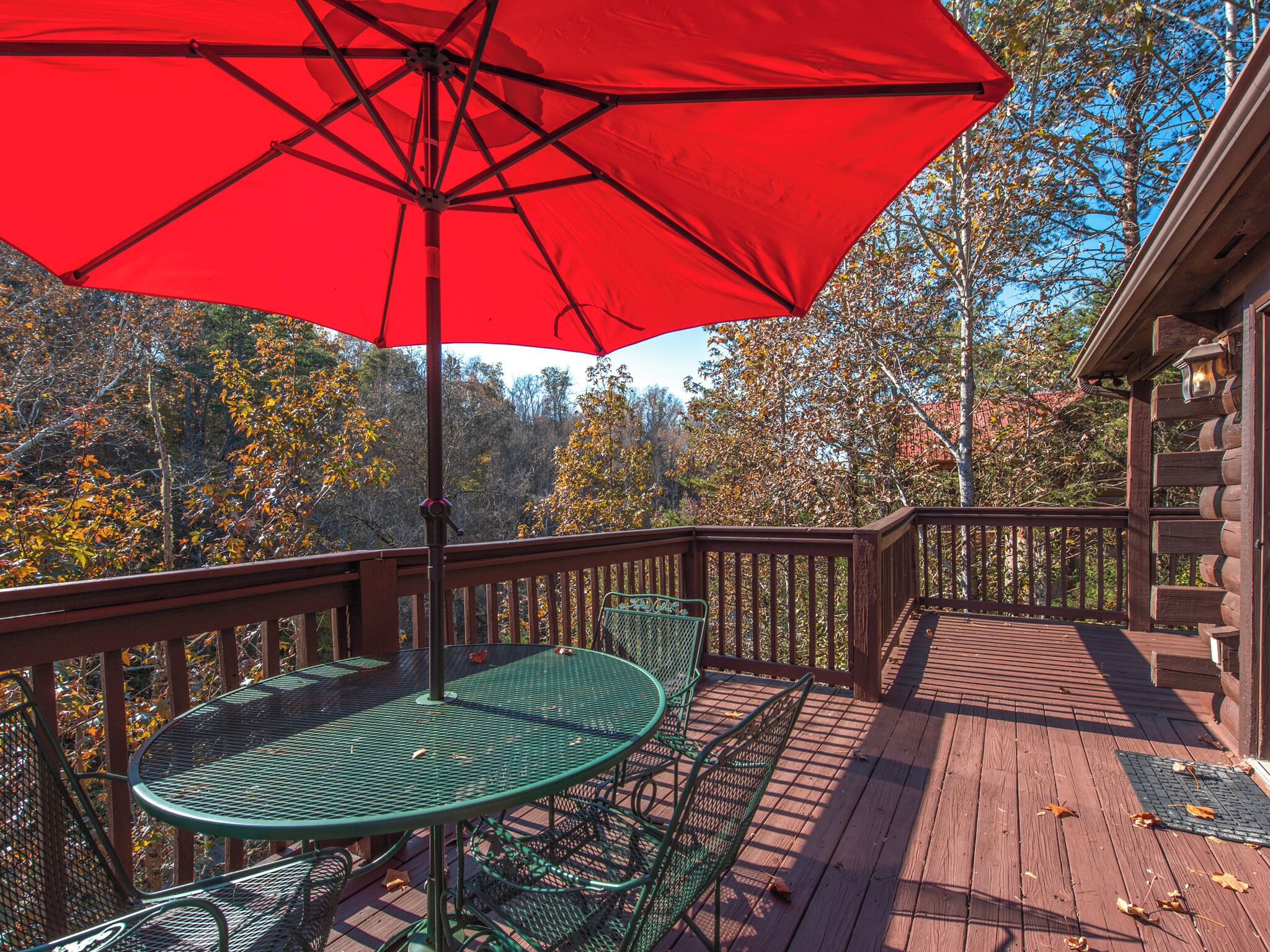 A wooden deck with a red umbrella, green metal table and chairs, and a view of trees and a building in the background.