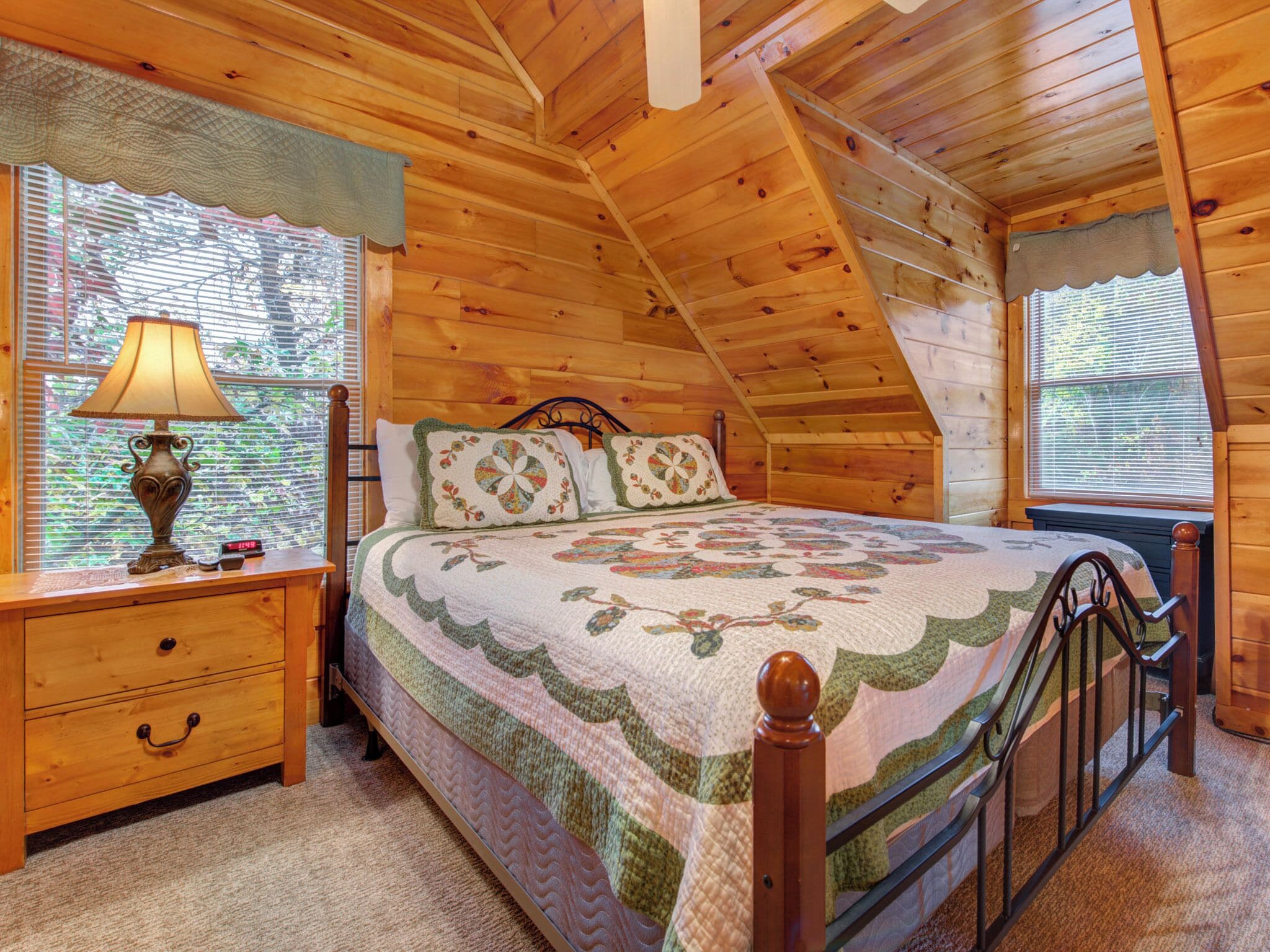 A cozy bedroom with a wooden ceiling, a bed with floral bedding, a nightstand with a lamp, and a window with blinds.
